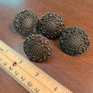 May include: Four antique bronze-colored cabinet knobs. Each knob has a round shape with a woven center and ornate, swirling details around the edges. The knobs are arranged on a wooden surface.