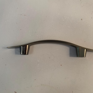 May include: A metallic cabinet pull with a smooth, arched design. The handle features a brushed silver finish on the top surface and two polished, cylindrical mounting posts extending downward from the ends of the curved bar.