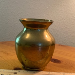 May include: A small, antique brass vase with a rounded body and a flared rim. The vase has a polished finish and is approximately 4 inches tall. The vase is sitting on a wooden surface.