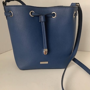 May include: A blue bucket bag with a drawstring closure and a shoulder strap. The bag has silver-tone hardware, including rings and a small rectangular emblem. The bag is made of a smooth, solid-colored material.