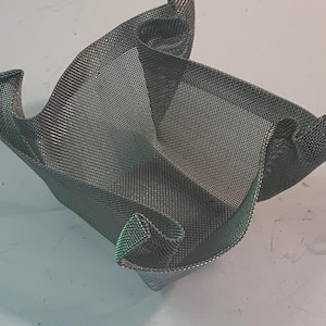 May include: A silver-colored, mesh basket with a square base and curved, folded edges. The basket has a textured appearance and is designed to hold small items. The mesh allows for air circulation and a modern aesthetic.