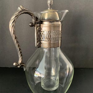 May include: A clear glass decanter with a silver-colored handle and accents. The decanter has a bulbous body and a cylindrical ice compartment. The lid is silver-colored and topped with a decorative finial. The handle has an ornate, textured design.