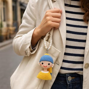 May include: A crochet doll, inspired by the painting "Girl with a Pearl Earring", with a blue hat, yellow top, and pearl necklace. The doll is attached to a white bag. The doll is handmade.