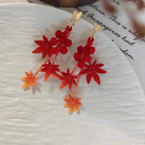 Peut inclure: Boucles d'oreilles pendantes en forme de feuilles rouges et oranges, avec des clous en forme de cœur dorés. Les boucles d'oreilles présentent des motifs de feuilles détaillés, parfaits pour une touche d'élégance inspirée de la nature.