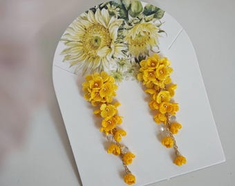 May include: A pair of bright yellow floral earrings. The earrings are made of small, crocheted flowers and hang from delicate gold chains. The earrings are displayed on a white card with a sunflower illustration.
