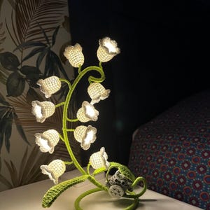 Könnte beinhalten: Eine gehäkelte Maiglöckchen-Lampe mit einer kleinen gehäkelten Eulenfigur an der Basis. Die Lampe ist eingeschaltet und die weißen Blüten sind beleuchtet.