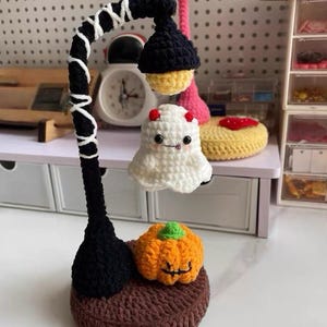 Op de afbeelding: Een handgemaakte Halloween-thema lamp gehaakt met een zwarte lantaarnpaal en een zwarte lampenkap met geel licht. Een wit spook met rode hoorns en een oranje pompoen zijn ook gehaakt. De lamp staat op een bruine gehaakte basis.