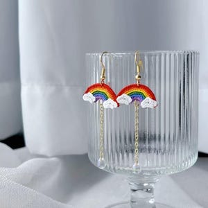 May include: A pair of rainbow-shaped earrings with gold-colored hooks and chains. The earrings feature a crocheted rainbow design with red, orange, yellow, green, blue, and purple colors. White cloud accents are at the base of each rainbow, and a clear crystal bead hangs from each chain.