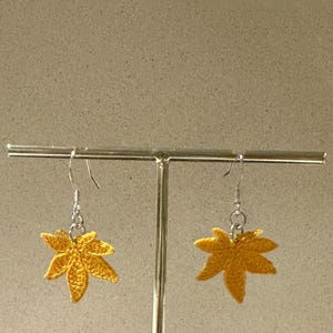May include: A pair of yellow crocheted maple leaf earrings hanging from silver hooks on a display stand.