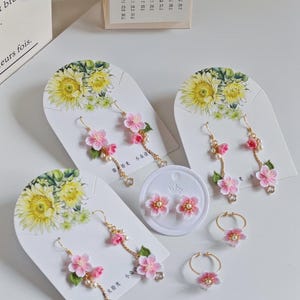 Peut inclure: Trois paires de boucles d'oreilles délicates en crochet avec des accents de perles et une bague avec une fleur en crochet. Les boucles d'oreilles sont présentées sur des cartes blanches avec des motifs floraux. La bague est sur une surface blanche.