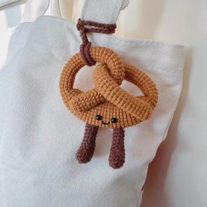 Può includere: Un ciondolo a forma di pretzel all'uncinetto con un volto sorridente e due gambe marroni, appeso a una borsa tote bianca. Il pretzel è di un caldo colore marrone dorato e le gambe sono di una tonalità di marrone più scura. Il ciondolo è attaccato alla borsa con un anello all'uncinetto marrone.