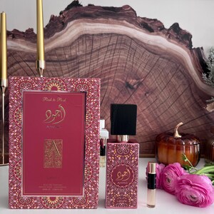 K&ouml;nnte beinhalten: Ein rosa Parf&uuml;mflakon und eine passende Schachtel mit goldenen Akzenten. Auf der Schachtel und der Flasche ist arabische Schrift und der Markenname "AIWAD" zu sehen. Ein kleines Probenfl&auml;schchen und rosa Blumen sind ebenfalls vorhanden. Im Hintergrund ein Holzkunstwerk.