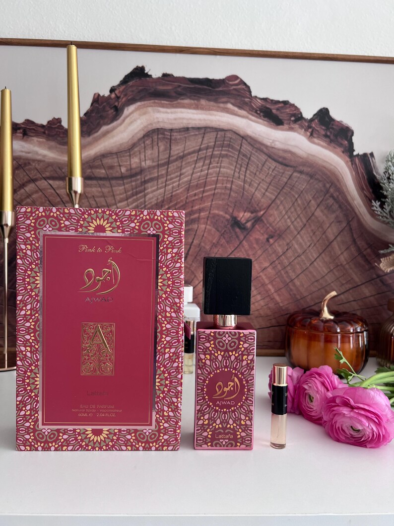 K&ouml;nnte beinhalten: Ein rosa Parf&uuml;mflakon und eine passende Schachtel mit goldenen Akzenten. Auf der Schachtel und der Flasche ist arabische Schrift und der Markenname "AIWAD" zu sehen. Ein kleines Probenfl&auml;schchen und rosa Blumen sind ebenfalls vorhanden. Im Hintergrund ein Holzkunstwerk.