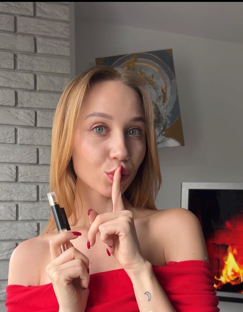 K&ouml;nnte beinhalten: Eine Frau in einem roten schulterfreien Oberteil h&auml;lt eine schwarze und transparente Lippenstift-Tube. Sie deutet mit einem Finger auf ihre Lippen und signalisiert Schweigen. Ein kleines Halbmond-Tattoo ist auf ihrem Arm zu sehen.