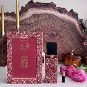 K&ouml;nnte beinhalten: Ein rosa Parf&uuml;mflaschen- und Box-Set mit einem goldenen Muster. Auf der Box steht "Pink to Pink" und arabische Schriftzeichen. Die Flasche hat einen schwarzen Deckel und einen goldenen Hals. Eine kleine Probe und rosa Blumen sind ebenfalls vorhanden.