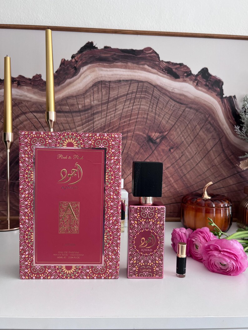 K&ouml;nnte beinhalten: Ein rosa Parf&uuml;mflaschen- und Box-Set mit einem goldenen Muster. Auf der Box steht "Pink to Pink" und arabische Schriftzeichen. Die Flasche hat einen schwarzen Deckel und einen goldenen Hals. Eine kleine Probe und rosa Blumen sind ebenfalls vorhanden.