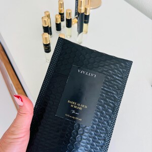 K&ouml;nnte beinhalten: Schwarzer Luftpolsterumschlag mit einem Etikett mit der Aufschrift "LATTAFA" und "Badee Al Oud Sublime For men". Im Hintergrund befinden sich mehrere kleine Parf&uuml;mflaschen mit goldenen Kappen und schwarzen Etiketten. Die Flaschen stehen auf einer wei&szlig;en Oberfl&auml;che.