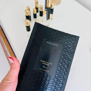 K&ouml;nnte beinhalten: Schwarzer Luftpolsterumschlag mit einem Etikett mit der Aufschrift "Badee Al Oud Sublime For men". Im Hintergrund befinden sich mehrere kleine Parf&uuml;mflaschen mit goldenen Kappen. Der Umschlag enth&auml;lt 10 ml Parf&uuml;m.