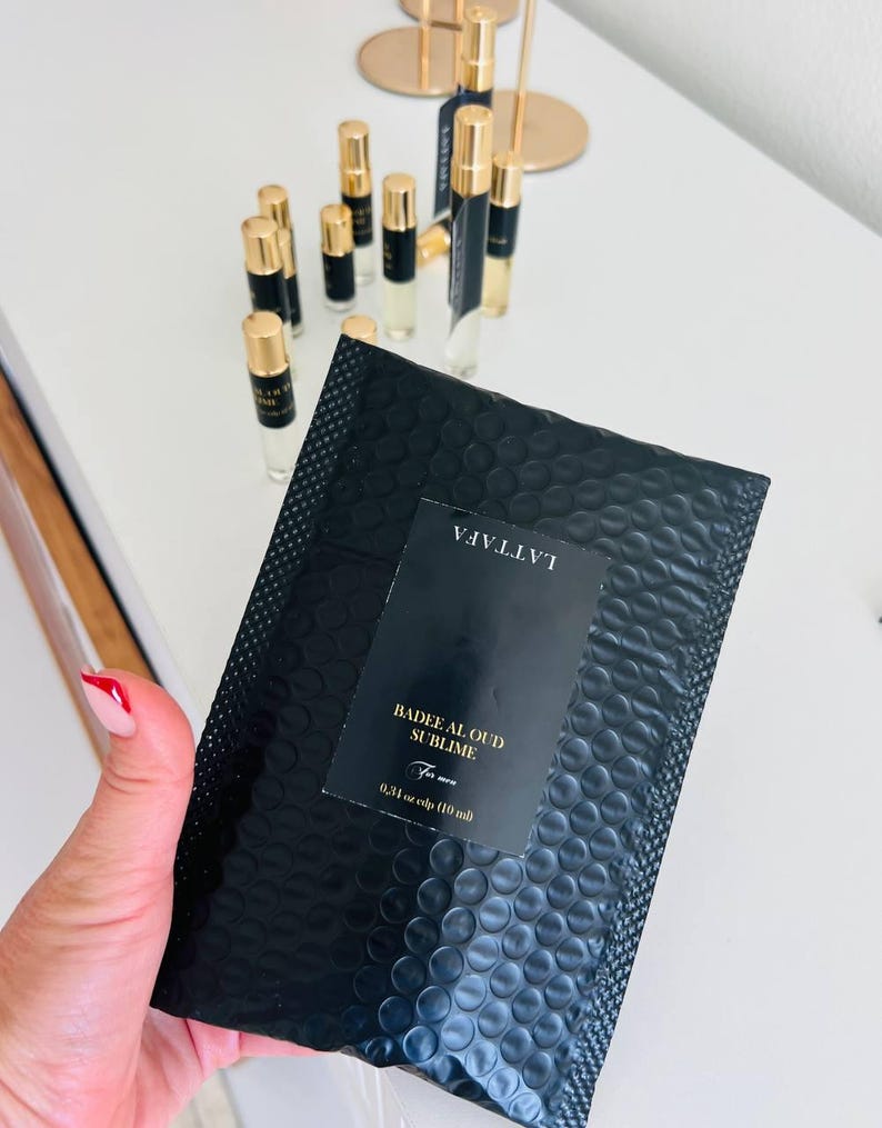 K&ouml;nnte beinhalten: Schwarzer Luftpolsterumschlag mit einem Etikett mit der Aufschrift "Badee Al Oud Sublime For men". Im Hintergrund befinden sich mehrere kleine Parf&uuml;mflaschen mit goldenen Kappen. Der Umschlag enth&auml;lt 10 ml Parf&uuml;m.