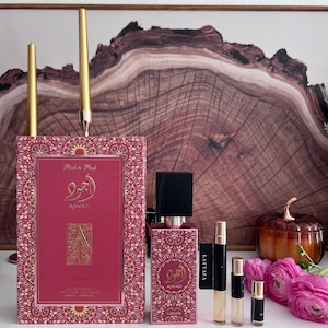 K&ouml;nnte beinhalten: Eine Pr&auml;sentation von rosa Parf&uuml;mflaschen und einer Schachtel mit goldenen Akzenten. Die Schachtel und die Flasche tragen arabische Schriftzeichen und den Text "Pink to Pink" und "AJWAD". Mehrere kleine Probeflaschen und rosa Blumen sind ebenfalls vorhanden, mit zwei goldenen Kerzen im Hintergrund.