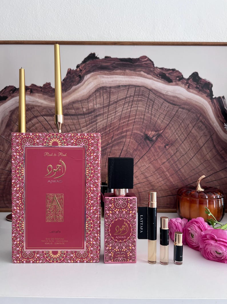 K&ouml;nnte beinhalten: Eine Pr&auml;sentation von rosa Parf&uuml;mflaschen und einer Schachtel mit goldenen Akzenten. Die Schachtel und die Flasche tragen arabische Schriftzeichen und den Text "Pink to Pink" und "AJWAD". Mehrere kleine Probeflaschen und rosa Blumen sind ebenfalls vorhanden, mit zwei goldenen Kerzen im Hintergrund.