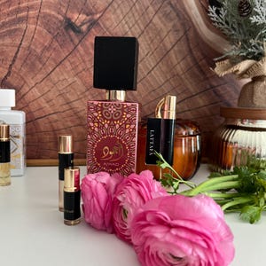 K&ouml;nnte beinhalten: Eine Anordnung von Parf&uuml;mflaschen und Probenfl&auml;schchen, darunter eine gro&szlig;e Flasche mit schwarzem Deckel und goldenen und roten Verzierungen. Kleine Fl&auml;schchen und rosa Blumen sind im Vordergrund angeordnet.