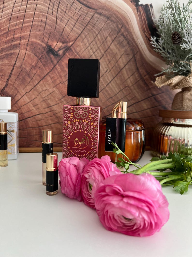 K&ouml;nnte beinhalten: Eine Anordnung von Parf&uuml;mflaschen und Probenfl&auml;schchen, darunter eine gro&szlig;e Flasche mit schwarzem Deckel und goldenen und roten Verzierungen. Kleine Fl&auml;schchen und rosa Blumen sind im Vordergrund angeordnet.