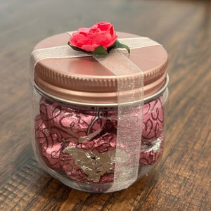 Op de afbeelding: Een doorzichtige pot gevuld met roze folie-verpakte snoepjes. De pot heeft een roze deksel, een zilveren lint en een kleine roze roos bovenop. De snoepjes zijn zichtbaar door de transparante zijkanten van de pot.