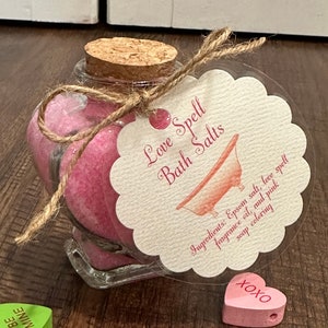 May include: A glass jar filled with pink bath salts, tied with twine and a heart-shaped tag that reads "Love Spell Bath Salts". The tag also includes a list of ingredients: Epsom salt, love spell fragrance oil, and pink soap coloring. The jar is surrounded by small wooden hearts with various messages, including "Be Mine", "Only You", and "XOXO".