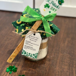 Può includere: Un barattolo di vetro pieno di ingredienti per un mix di biscotti, decorato con tessuto verde e oro e un nastro verde. Un cucchiaio di legno con una spatola bianca attaccata è legato al barattolo con uno spago. La spatola ha il testo "St. Patrick's Day". Un'etichetta attaccata al barattolo recita "St. Patrick's Day M & M Cookie Mix in a Jar".