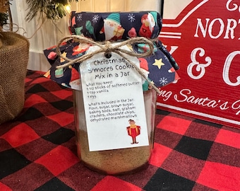 May include: A glass jar filled with cookie mix, labeled "Christmas S'mores Cookie Mix in a Jar." The jar is adorned with a fabric lid featuring festive gnome and star patterns, tied with twine. The jar sits on a red and black plaid surface.