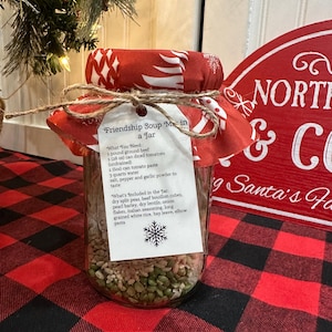 Könnte beinhalten: Ein klares Glas mit getrockneten Suppenzutaten, bedeckt mit einem roten Stoffdeckel und mit Bindfaden gebunden. Ein Etikett trägt die Aufschrift "Friendship Soup Mix in a Jar" mit einer Zutatenliste und einer Schneeflocke. Das Glas steht auf einer rot-schwarz karierten Oberfläche.