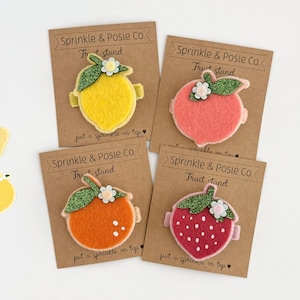 May include: Four felt fruit hair clips: a yellow lemon, a pink peach, an orange, and a red strawberry. Each clip is attached to a brown card with the text "Sprinkle & Posie Co. Fruit stand". The fruit designs have glittery green leaves and small white flowers.