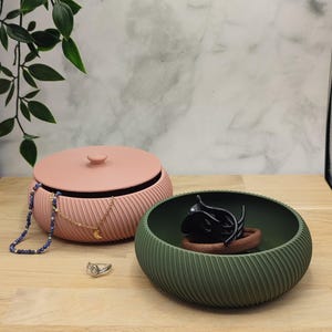 Könnte beinhalten: Zwei dekorative Schalen und Schmuck auf einer Holzoberfläche. Eine rosa Schale mit Deckel und eine grüne Schale mit einem schwarzen Objekt im Inneren. Eine blaue Perlenkette, eine goldene Halskette mit Mondanhänger und ein Ring sind ebenfalls vorhanden.