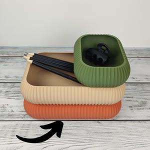 Könnte beinhalten: Ein Stapel aus drei rechteckigen, gerippten Silikonschalen in Orange, Beige und Olivgrün. Die oberste olivgrüne Schale enthält eine schwarze Haarspange, während die beige Schale vier schwarze Bleistifte enthält. Ein schwarzer Pfeil zeigt auf die untere Schale.