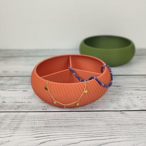 Könnte beinhalten: Eine terrakottafarbene, runde, gerippte Aufbewahrungsschale mit drei Innenfächern. Eine goldfarbene Kette mit kleinen runden Anhängern und eine blaue Perlenkette liegen über dem Rand der Schale. Dahinter befindet sich eine schlichte olivgrüne Schale.