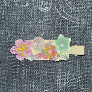 Puede incluir: Una pinza para el pelo decorativa con una flor rosa, un gato blanco, una estrella y adornos coloridos. La pinza es de color beige claro y tiene un cierre de tipo horquilla. Los accesorios son de plástico y están adheridos a la pinza.
