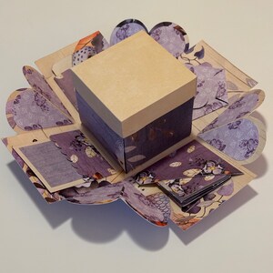 May include: A purple and brown explosion box with a beige lid. The box is decorated with floral patterns and has a small, square window on the front.