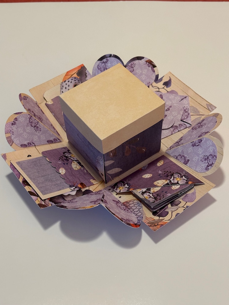May include: A purple and brown explosion box with a beige lid. The box is decorated with floral patterns and has a small, square window on the front.