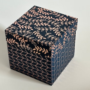 May include: A square gift box with a black and gold geometric pattern. The top of the box has a floral design with gold leaves on a black background.