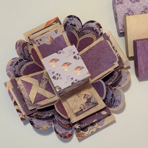 May include: A purple and brown explosion box with a floral pattern. The box is open and has multiple layers. The box is decorated with hearts and leaves.