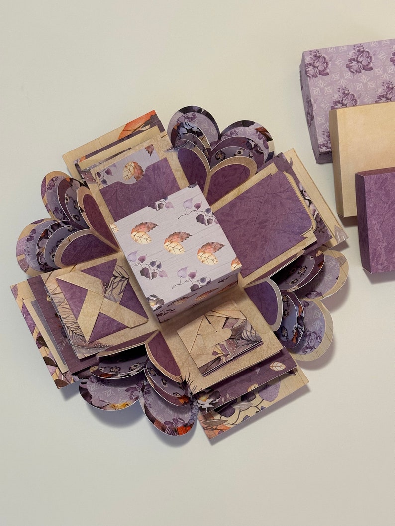 May include: A purple and brown explosion box with a floral pattern. The box is open and has multiple layers. The box is decorated with hearts and leaves.