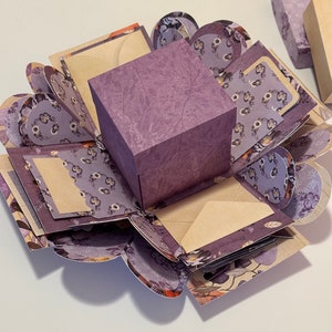 May include: A purple explosion box with a floral pattern. The box is open and has multiple layers, each with a different design. The box is perfect for gifts or special occasions.