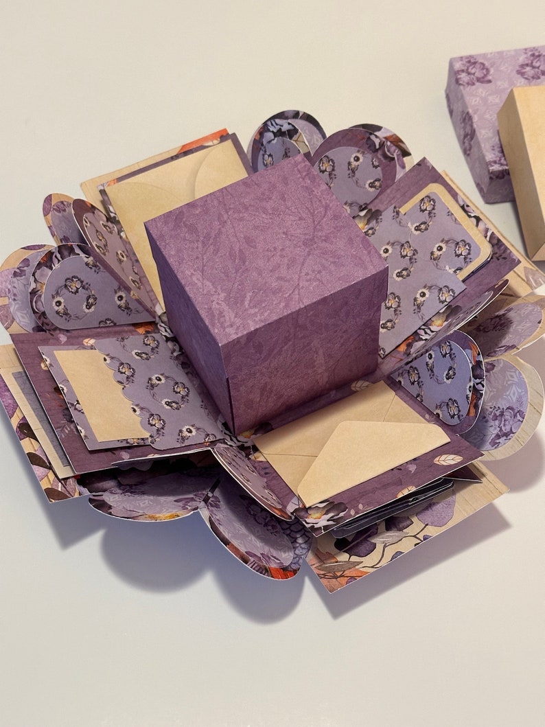 May include: A purple explosion box with a floral pattern. The box is open and has multiple layers, each with a different design. The box is perfect for gifts or special occasions.