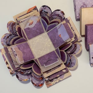 May include: A purple and cream explosion box with a floral pattern. The box is open and the inside is visible. The box is made of paper and has a delicate design.