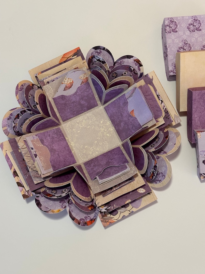 May include: A purple and cream explosion box with a floral pattern. The box is open and the inside is visible. The box is made of paper and has a delicate design.