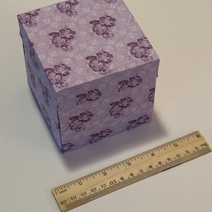 May include: A small, square, purple gift box with a floral pattern. The box is made of cardboard and has a hinged lid. The box is sitting on a white surface.