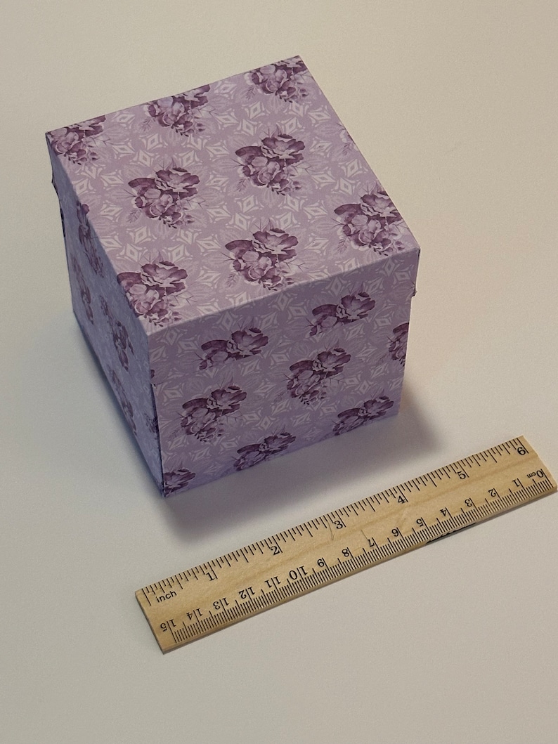 May include: A small, square, purple gift box with a floral pattern. The box is made of cardboard and has a hinged lid. The box is sitting on a white surface.