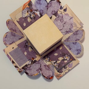 May include: A purple and cream explosion box with a floral pattern. The box is open and has a small square box inside.