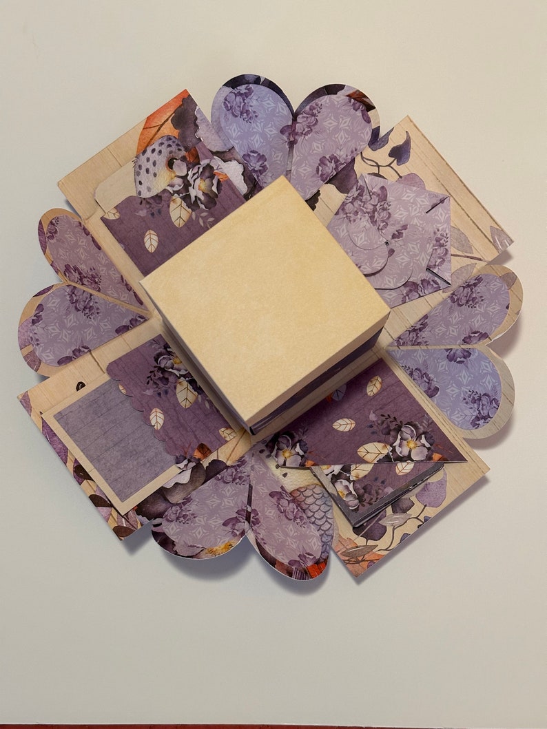 May include: A purple and cream explosion box with a floral pattern. The box is open and has a small square box inside.